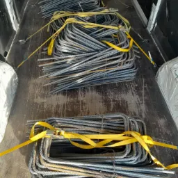Metalowe pręty zbrojeniowe, uformowane w kształt prostokątnych strzemion, zabezpieczone żółtymi pasami transportowymi, ułożone w kilku pakietach w przestrzeni ładunkowej białego busa.