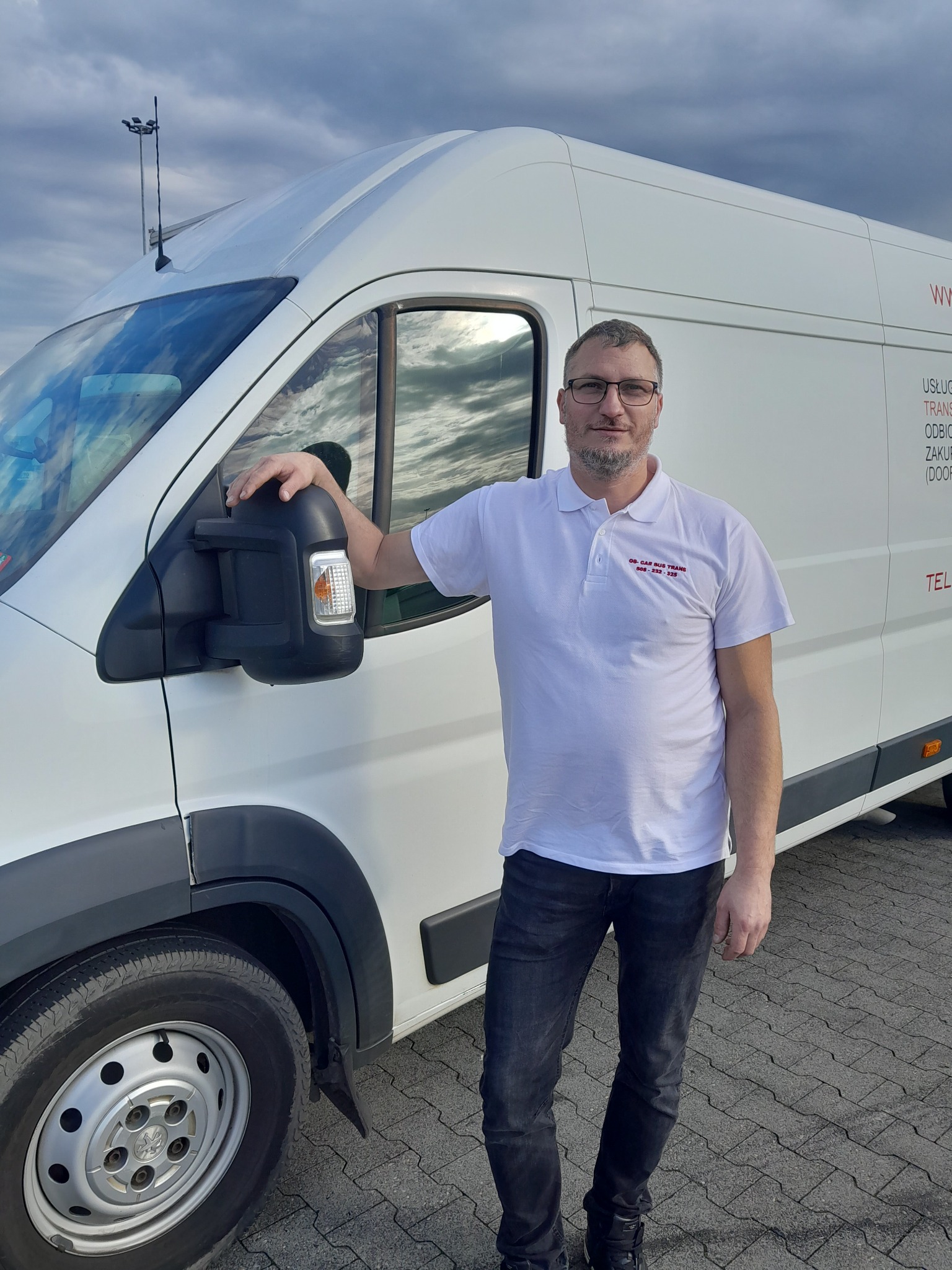Mężczyzna w okularach, opierający się o biały samochód dostawczy z widocznymi danymi kontaktowymi firmy i częściowo widocznym logo na koszulce, na tle pochmurnego nieba.