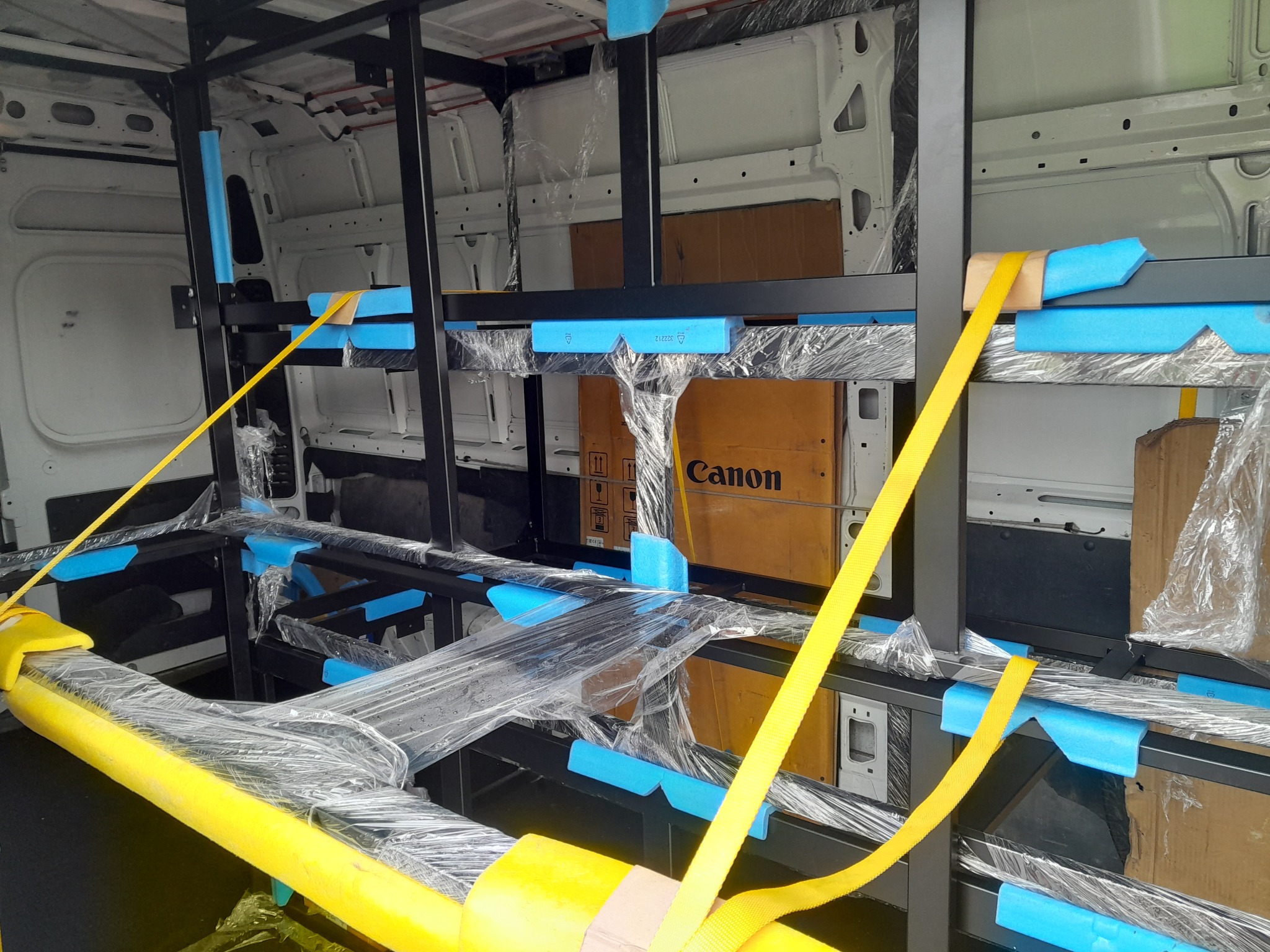 Wnętrze białego busa z zamontowaną, czarną metalową konstrukcją zabezpieczoną niebieskimi piankami i żółtymi pasami transportowymi, owiniętą folią stretch, przygotowaną do transportu. Widoczne...