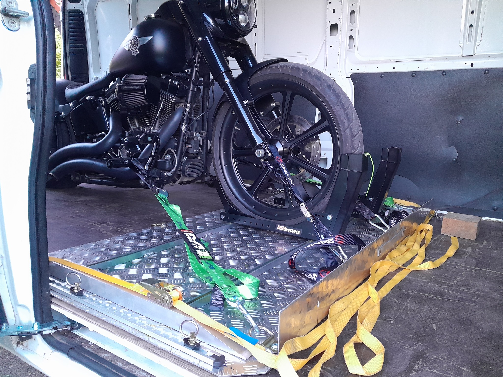 Czarny motocykl Harley-Davidson zabezpieczony pasami wewnątrz białego busa na metalowej platformie z rampą załadunkową.