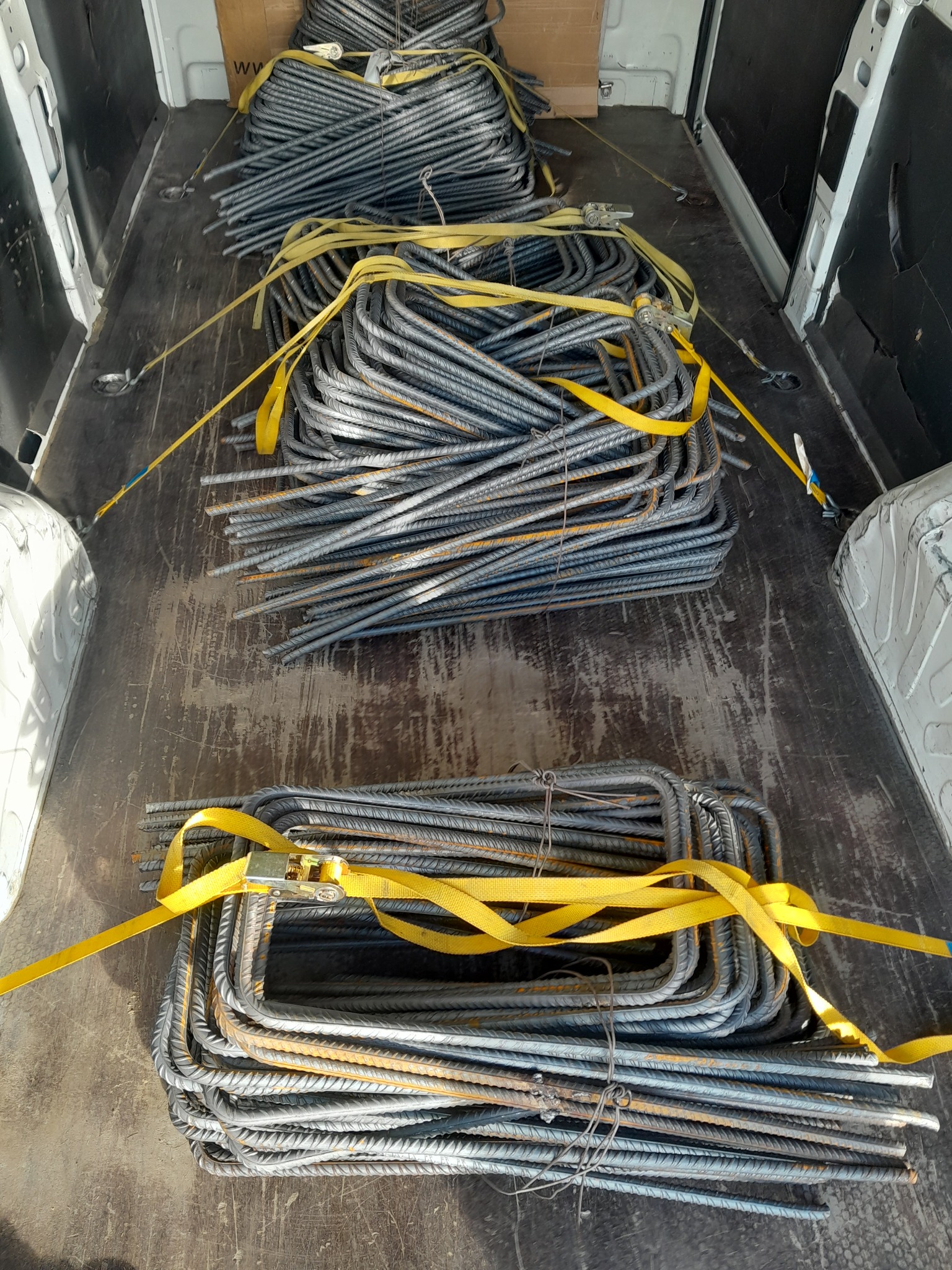 Metalowe pręty zbrojeniowe, uformowane w kształt prostokątnych strzemion, zabezpieczone żółtymi pasami transportowymi, ułożone w kilku pakietach w przestrzeni ładunkowej białego busa.