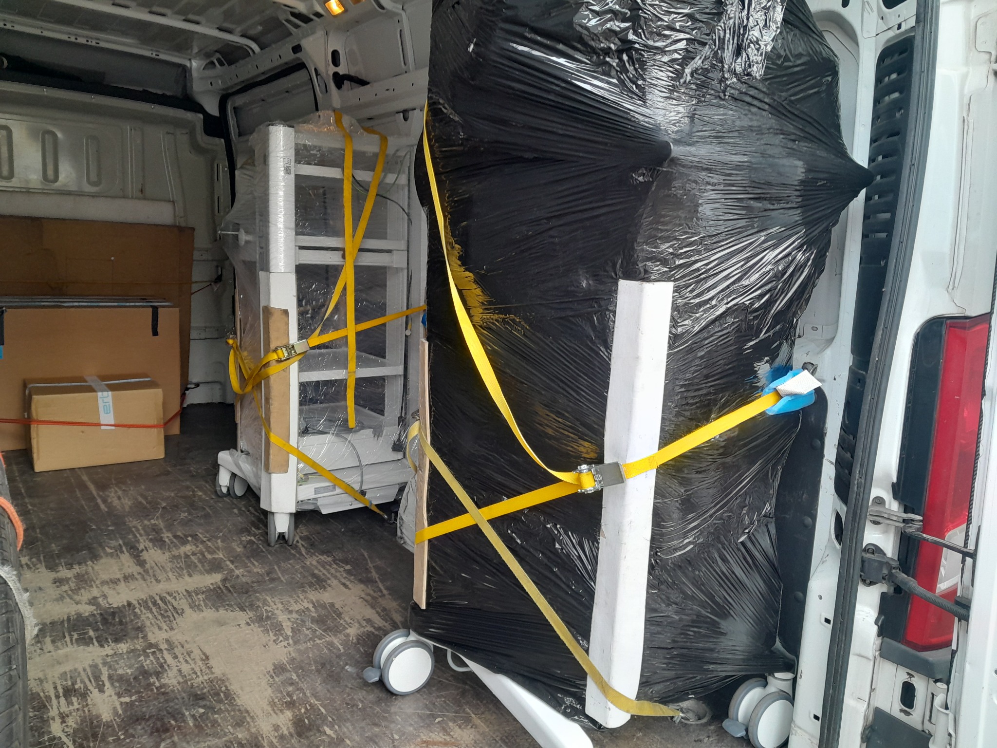 Wnętrze busa transportowego z zabezpieczonymi pasami transportowymi urządzeniami medycznymi: owiniętym czarną folią urządzeniem oraz wózkiem medycznym owiniętym folią stretch.