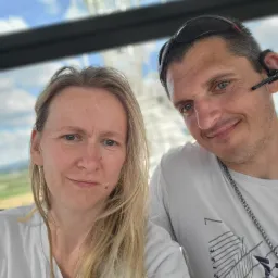 Selfie kobiety z blond włosami i mężczyzny z okularami na głowie, oboje uśmiechnięci na tle konstrukcji przypominającej maszt radiowy i błękitnego nieba.