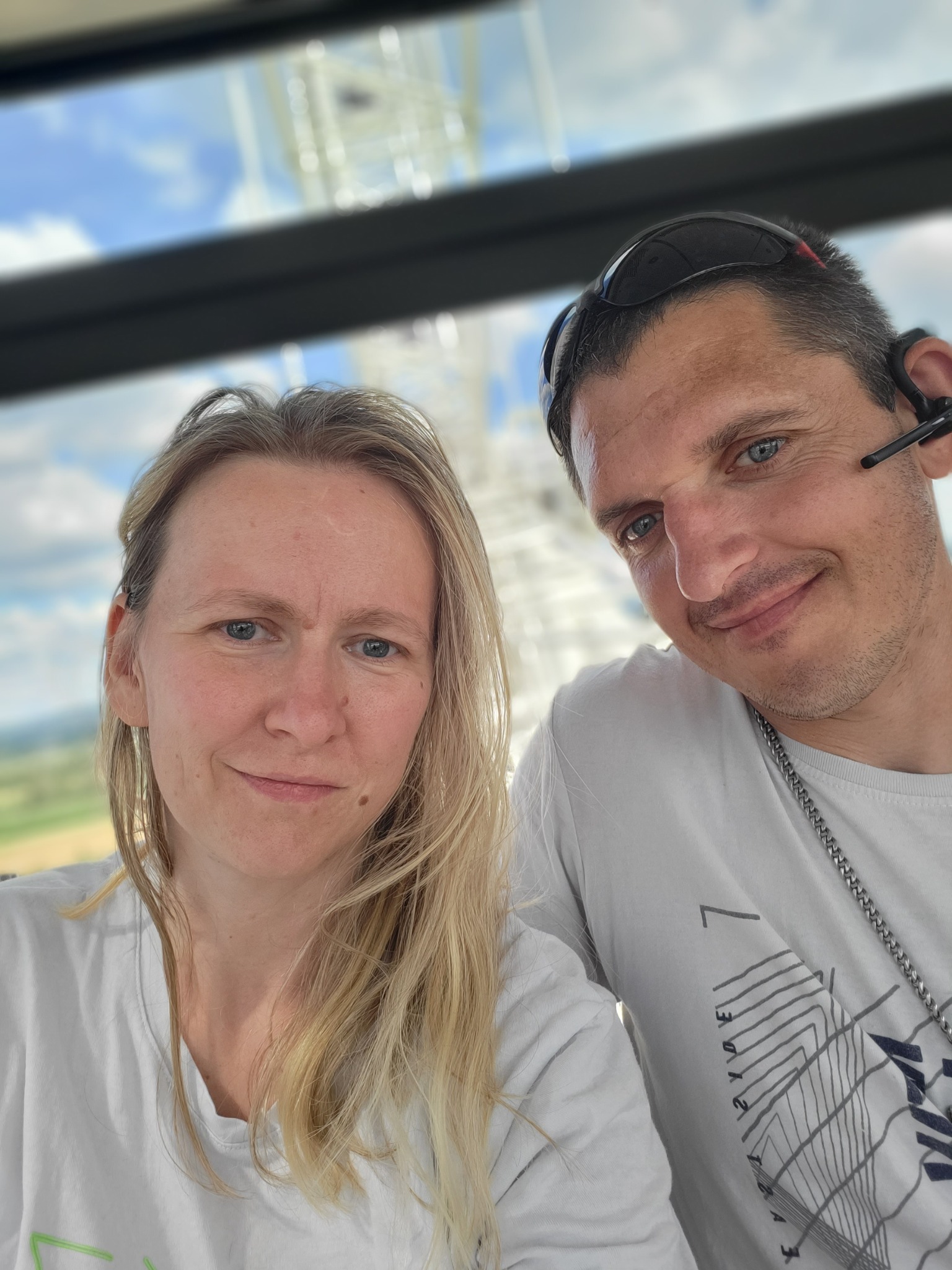 Selfie kobiety z blond włosami i mężczyzny z okularami na głowie, oboje uśmiechnięci na tle konstrukcji przypominającej maszt radiowy i błękitnego nieba.