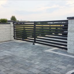 MarkoSpaw - Nowoczesna, przesuwna brama wjazdowa z poziomych, czarnych profili, zamontowana między murowanymi słupkami z szarego kamienia, na podjeździe z szarej kostki brukowej.