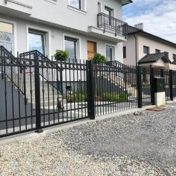 MarkoSpaw - Nowoczesne, czarne ogrodzenie metalowe z bramą, oddzielające posesję z dwoma budynkami mieszkalnymi, wysypaną szarą żwirową nawierzchnią.