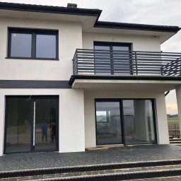 MarkoSpaw - Nowoczesny dom jednorodzinny z balkonem zabezpieczonym prostą, metalową balustradą z poziomych profili, duże przesuwne okna tarasowe i schody wejściowe wyłożone kostką.