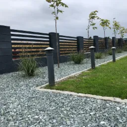 Nowoczesne ogrodzenie panelowe w kolorze grafitowym z podmur&oacute;wką, uzupełnione ozdobnym grysem i młodymi drzewkami, oświetlone niskimi lampami ogrodowymi.