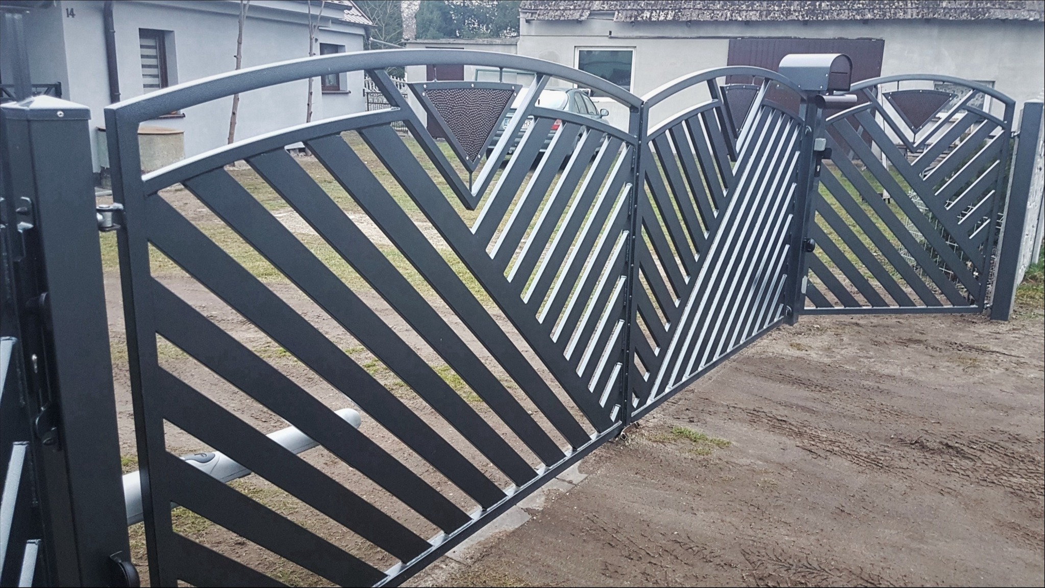 Nowoczesna, czarna brama wjazdowa z ukośnymi, białymi przerwami, wyposażona w automat, z widocznym numerem domu w tle.