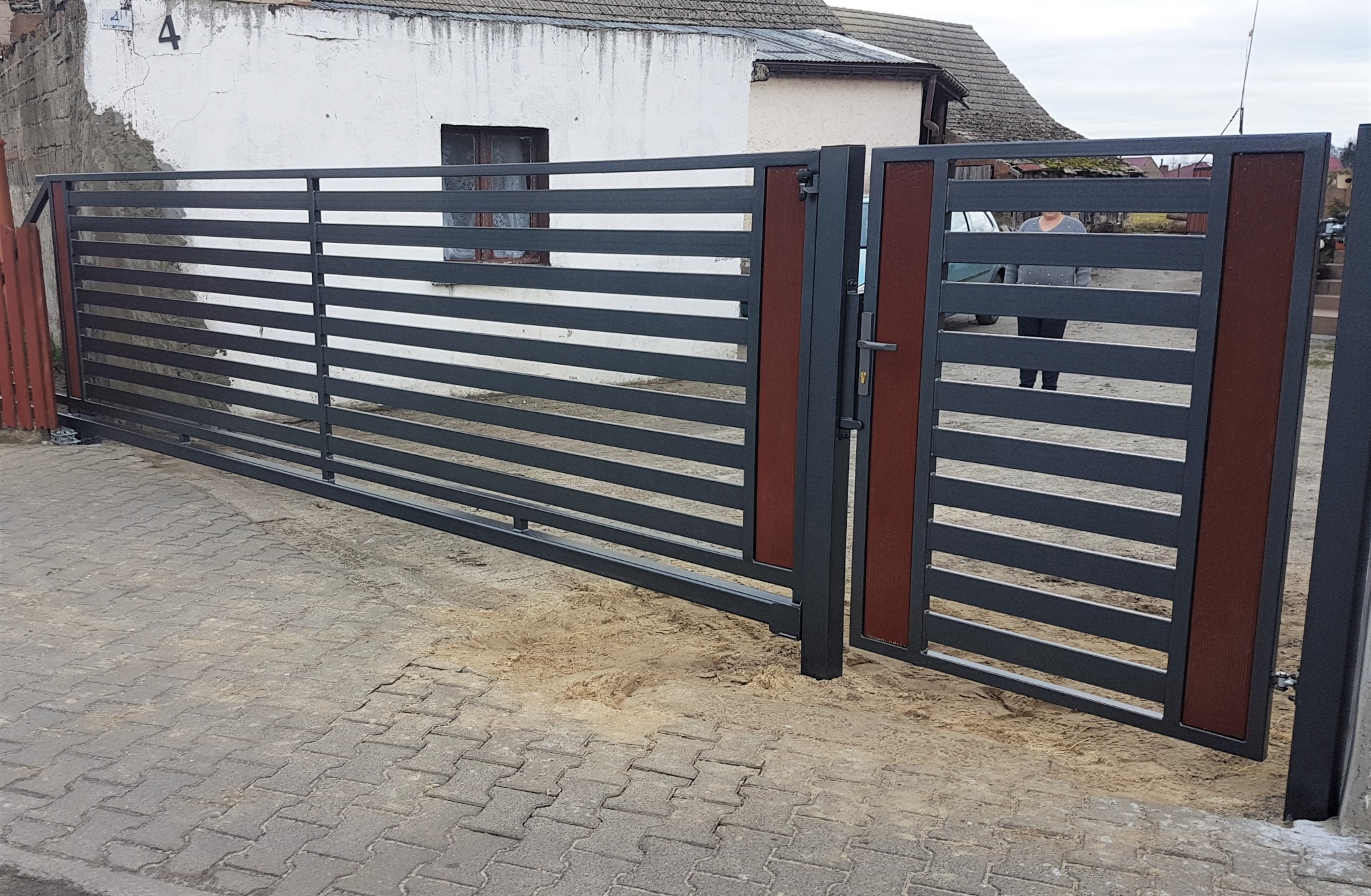 Nowoczesna brama wjazdowa przesuwna z poziomymi panelami w kolorze grafitowym i brązowymi akcentami, widoczna na tle kostki brukowej i budynku z numerem 4.