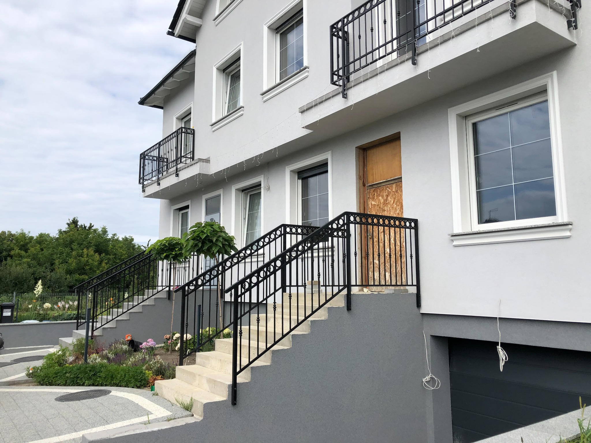 Elewacja domu z dwoma wejściami schodowymi z czarnymi, metalowymi balustradami, oraz balkony z podobnymi balustradami, drewniana płyta zabezpieczająca drzwi wejściowe.