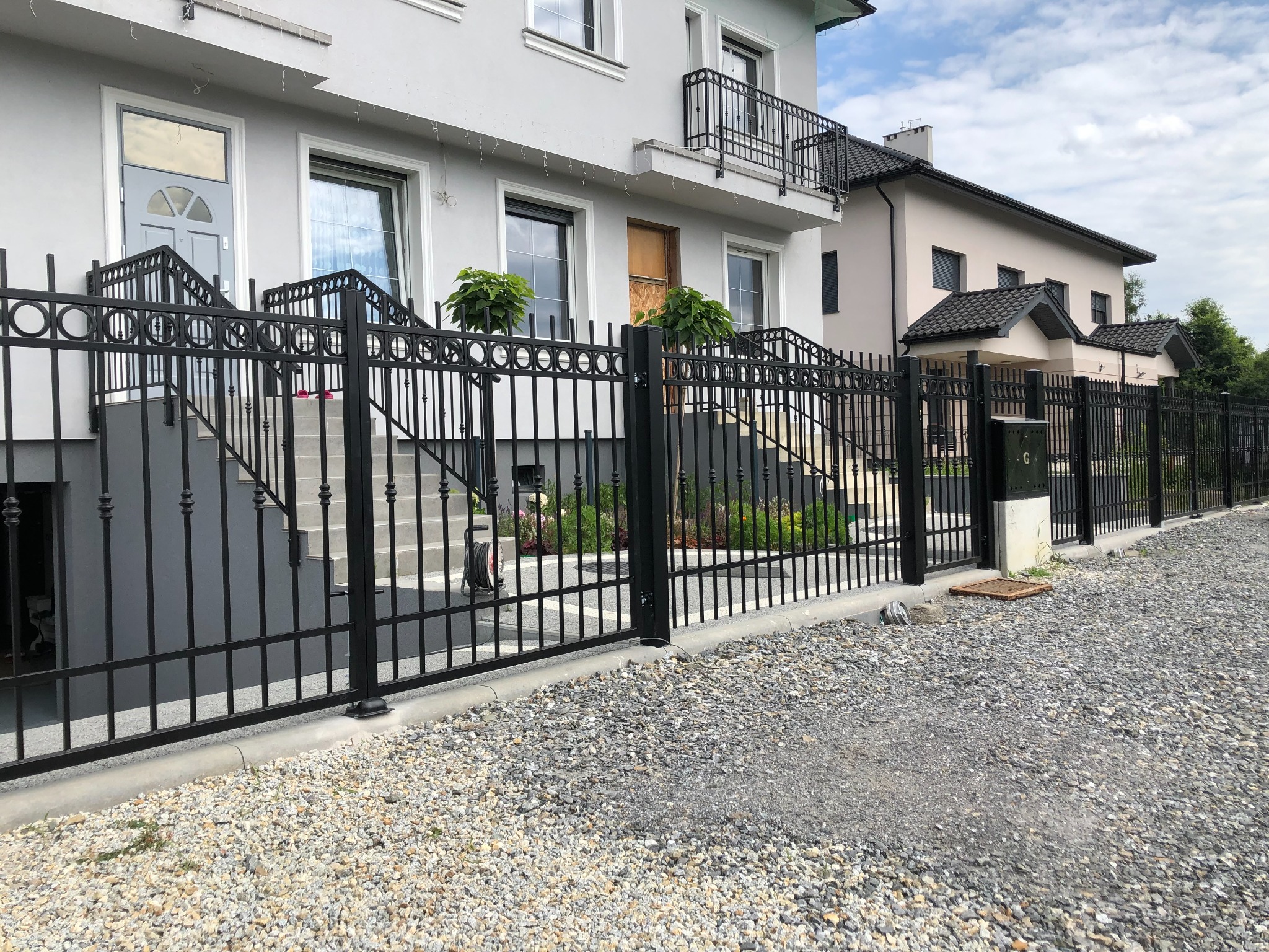 Nowoczesne, czarne ogrodzenie metalowe z bramą, oddzielające posesję z dwoma budynkami mieszkalnymi, wysypaną szarą żwirową nawierzchnią.