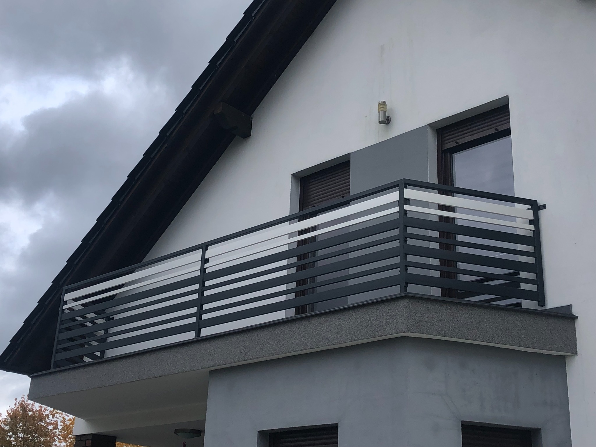Nowoczesna balustrada balkonowa z poziomych, czarnych profili metalowych, zamontowana na balkonie domu z szarą elewacją i brązowymi roletami.