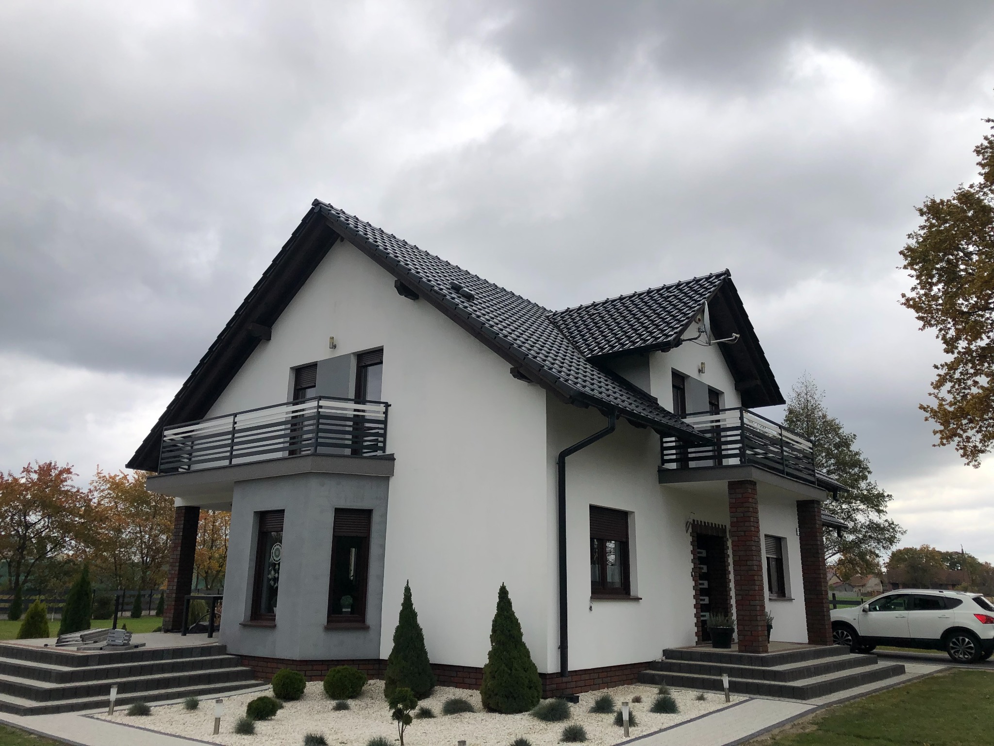 Nowoczesny dom jednorodzinny z balkonami z metalowymi balustradami, widok z zewnątrz, na pierwszym planie ozdobny ogródek z białym żwirem i małymi drzewkami.