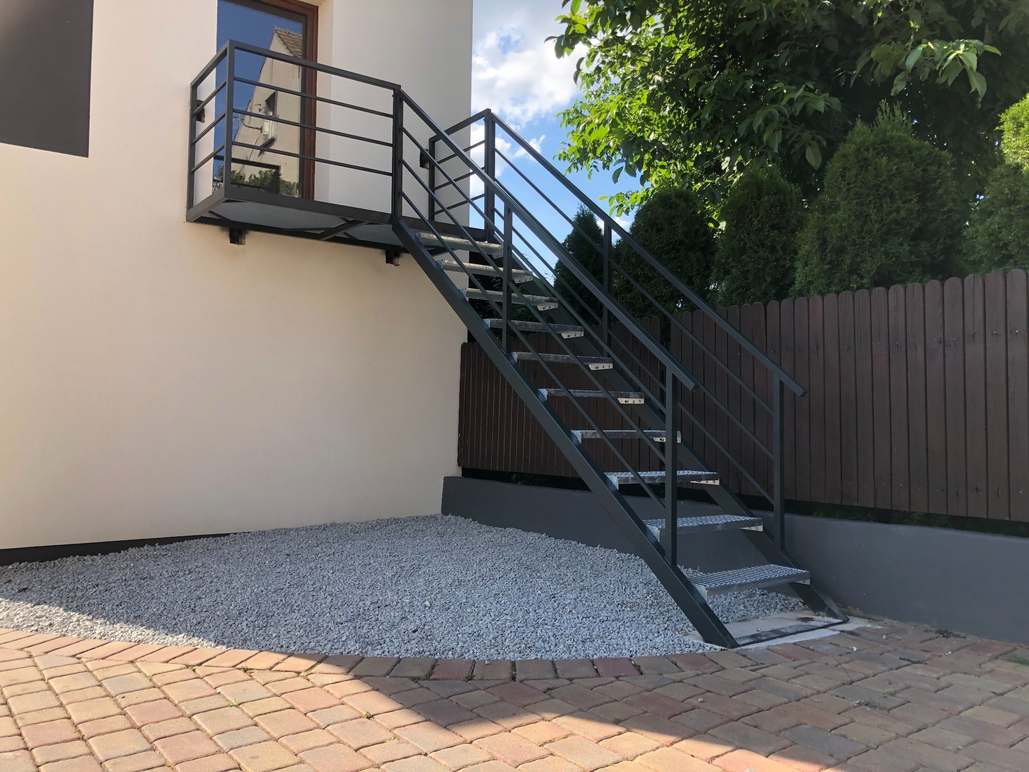 Zewnętrzne, metalowe schody prowadzące do balkonu przy jasnym budynku, z widoczną nawierzchnią z kostki brukowej i białego żwiru.