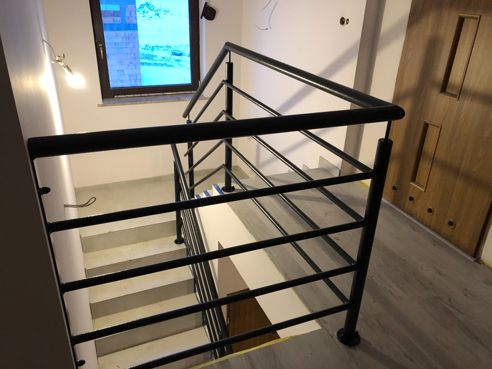 Wewnętrzne schody z czarną metalową balustradą, widok z góry, z oknem w tle i drewnianymi drzwiami po prawej stronie.