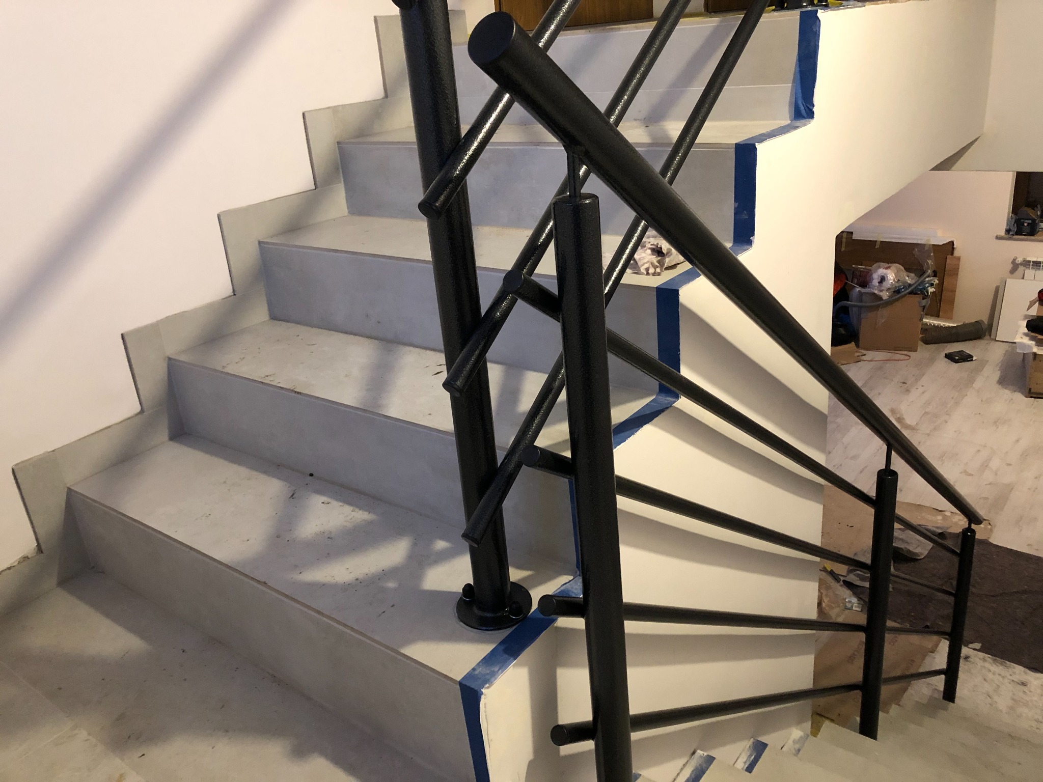Nowoczesne schody z metalową balustradą w trakcie montażu, zabezpieczone niebieską taśmą malarską, widoczne niedoskonałości wykończenia.