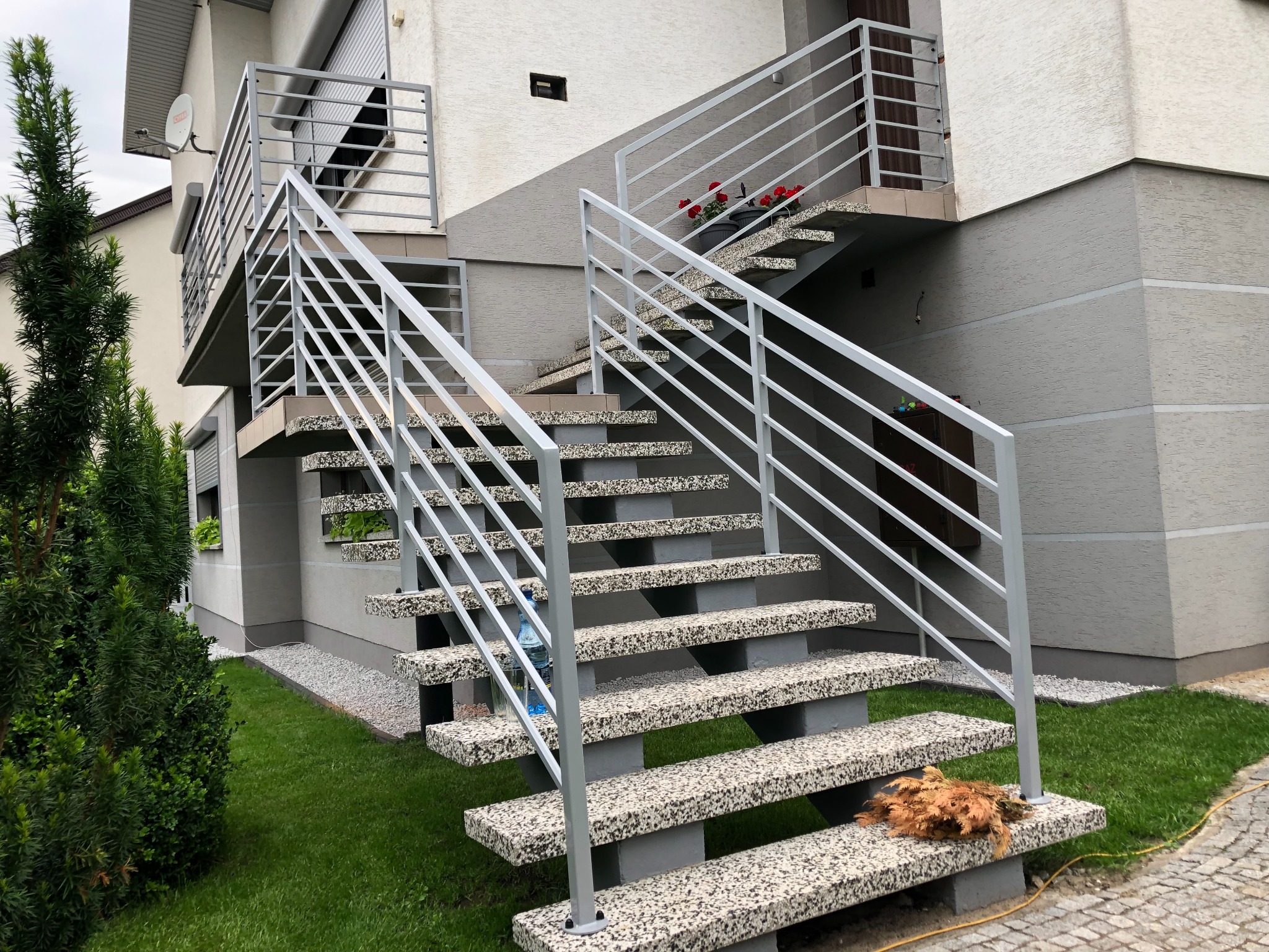 Zewnętrzne schody z szarymi metalowymi barierkami prowadzące na balkon, z dekoracyjnymi kwiatami w doniczkach na górnym poziomie. Na dolnym stopniu leżą uschnięte liście.