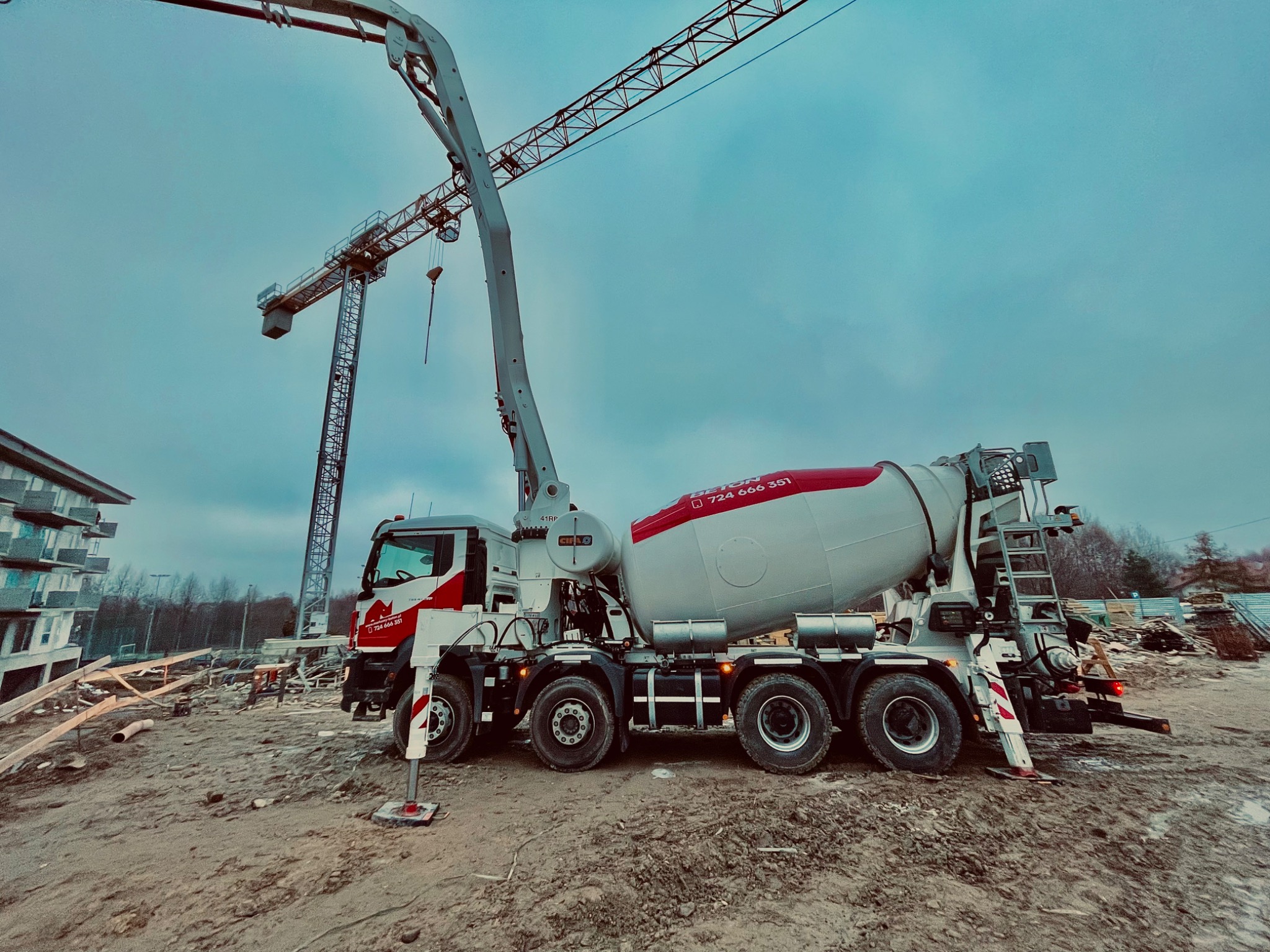 Ciężarówka z betonomieszarką i wysięgnikiem pompy stoi na placu budowy, w tle żuraw wieżowy i budynek w stanie surowym.