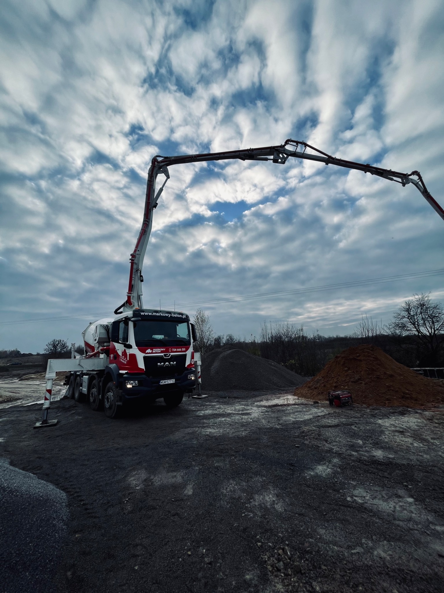 Ciężarówka z pompą do betonu na placu budowy, widoczne pryzmy piasku i żwiru, pochmurne niebo w tle.