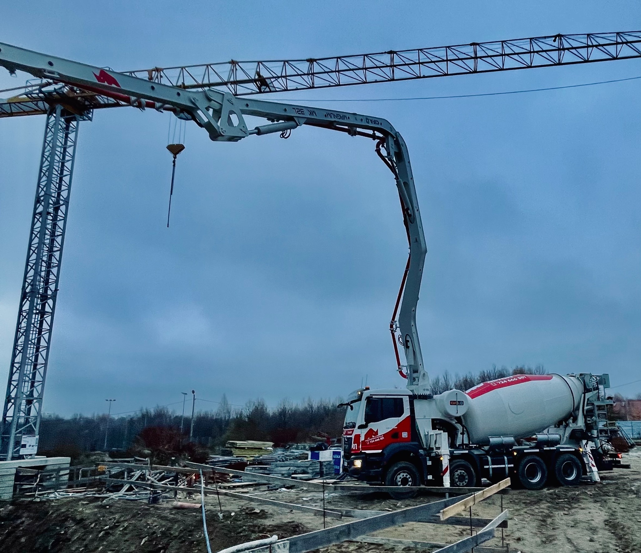 Betonomieszarka z pompą na budowie, widoczny żuraw budowlany i przygotowany szalunek fundamentów, pochmurne niebo.