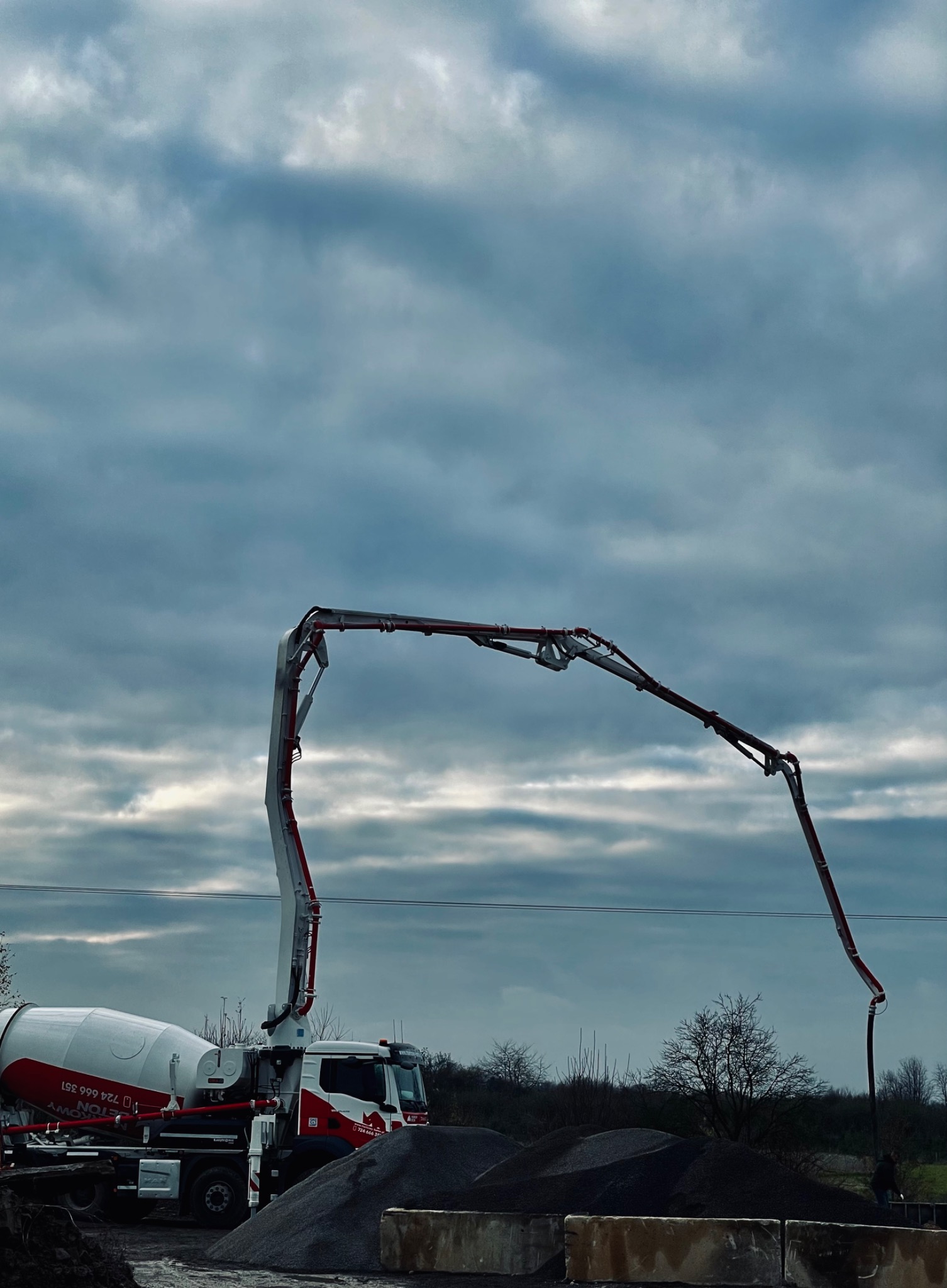 Pompa do betonu zamontowana na ciężarówce, z wysięgnikiem skierowanym w dół, w tle pochmurne niebo i hałda kruszywa.