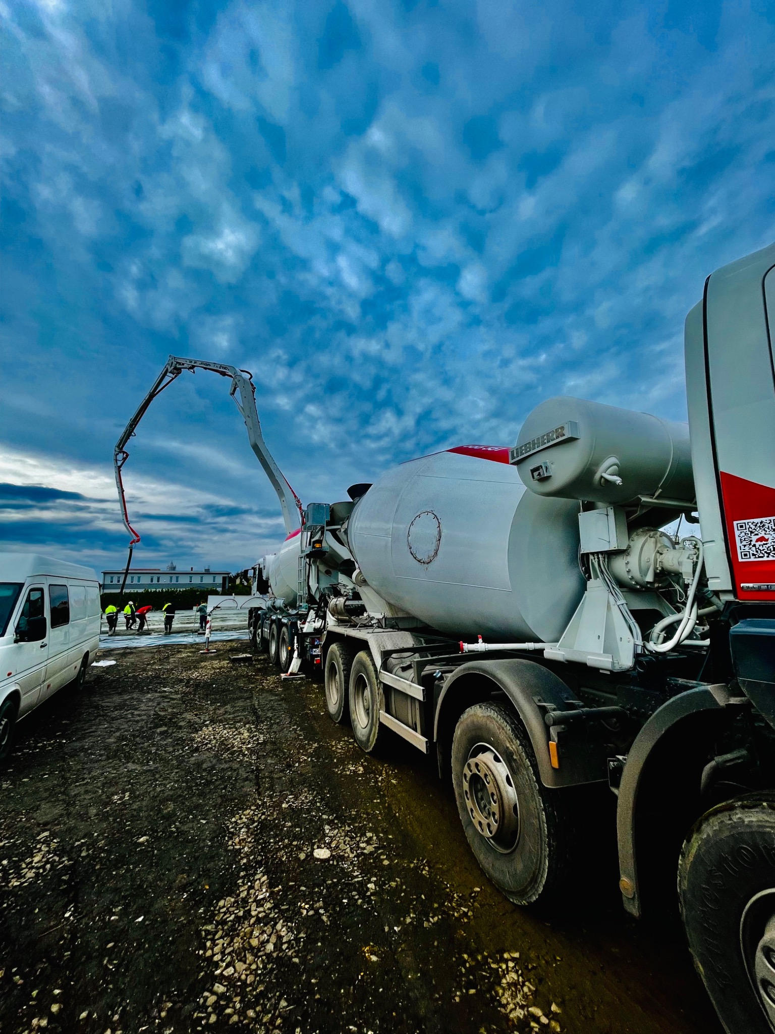 Ciężarówka z pompą do betonu na budowie, robotnicy w odblaskowych kamizelkach rozlewają beton, obok biały bus, pochmurne niebo.