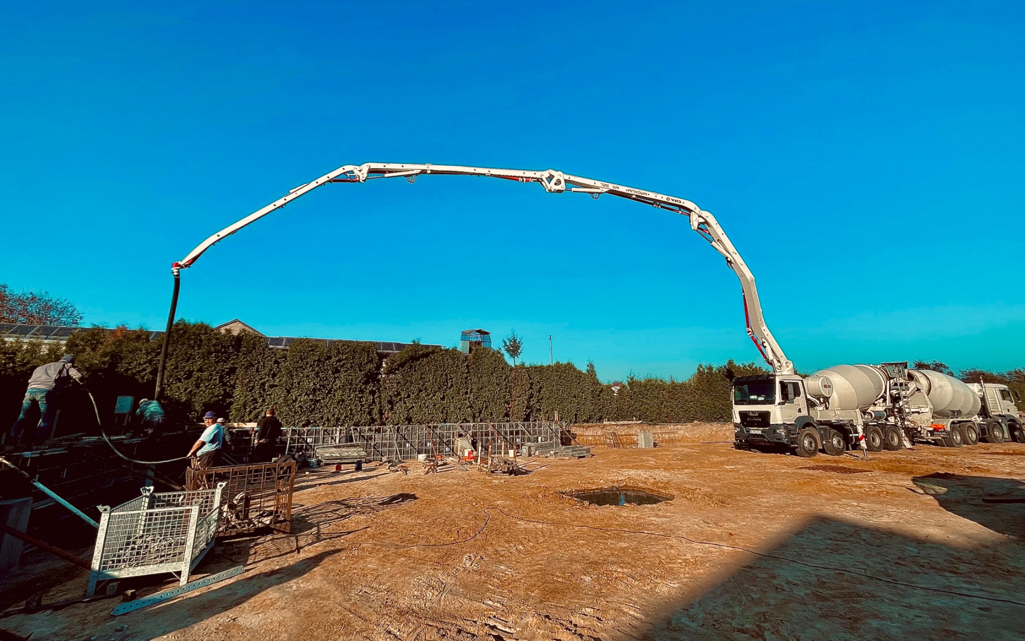 Betonomieszarka z długim ramieniem pompuje beton do szalunków fundamentowych na placu budowy, widoczni pracownicy w trakcie wylewania betonu.