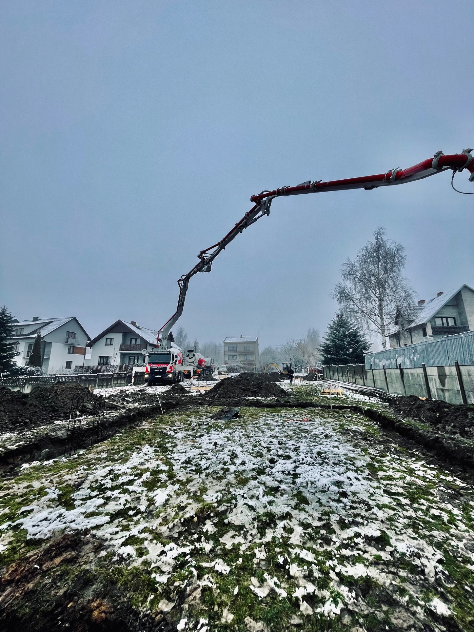 Pompa do betonu na budowie domu jednorodzinnego, z widoczną betonowozą w tle i częściowo zasypanymi śniegiem wykopami pod fundamenty.