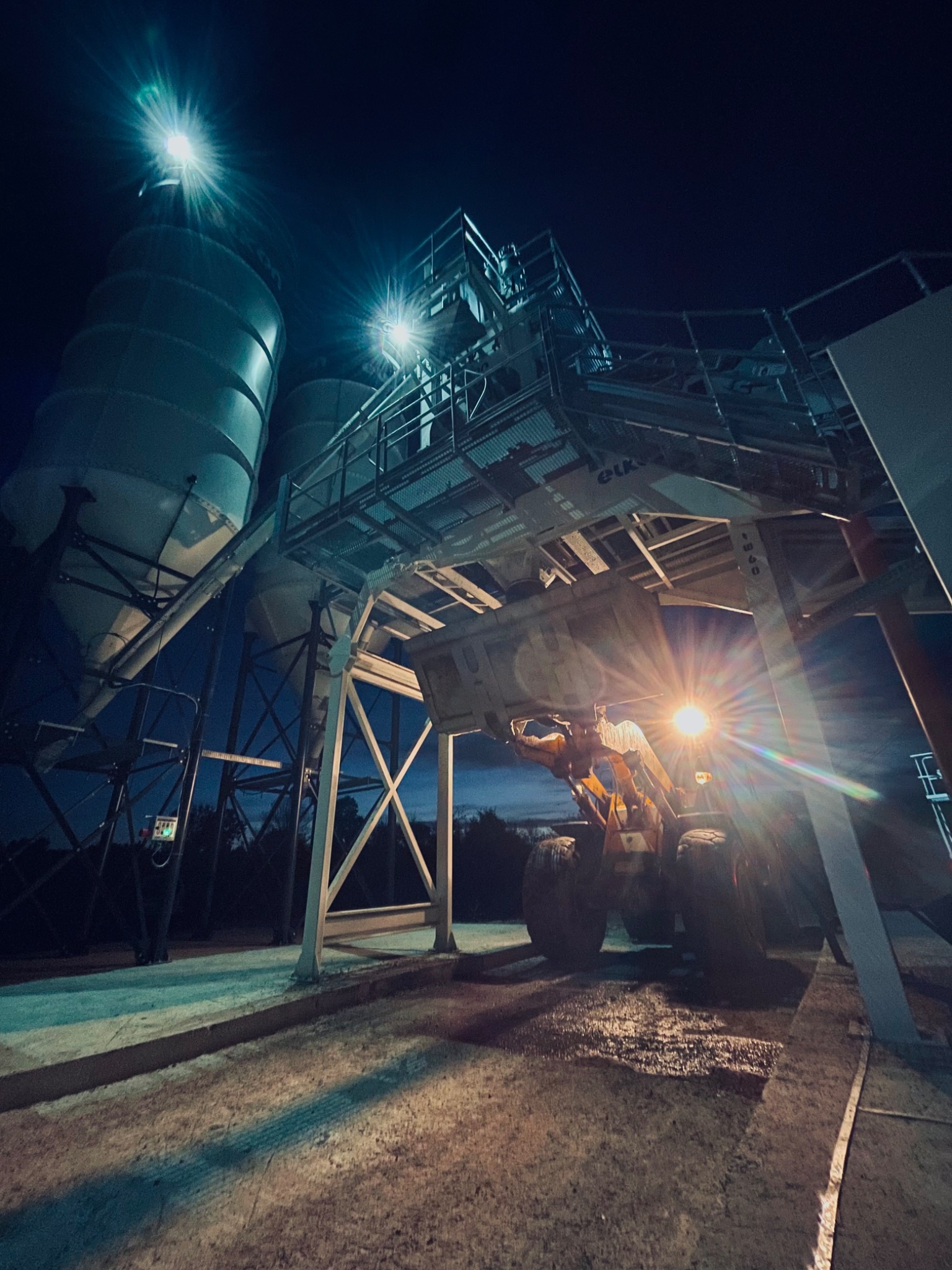 Nocny widok na betoniarnię z silosami i ładowarką teleskopową pod platformą, oświetloną jasnymi reflektorami.