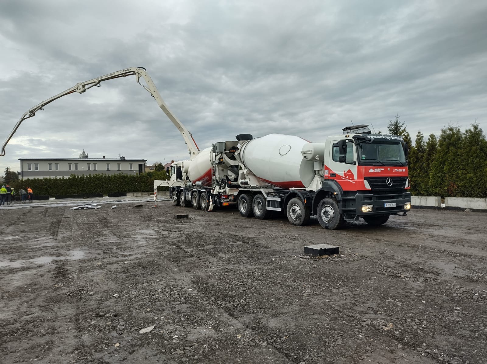 Dwa pojazdy betonomieszarki z pompą do betonu na placu budowy; widoczne fundamenty i grupa robotników w oddali.