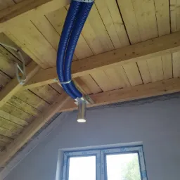 Dwie niebieskie rury wentylacyjne przymocowane do drewnianego stropu za pomocą metalowych obejm, z widoczną metalową puszką i oknem w tle.