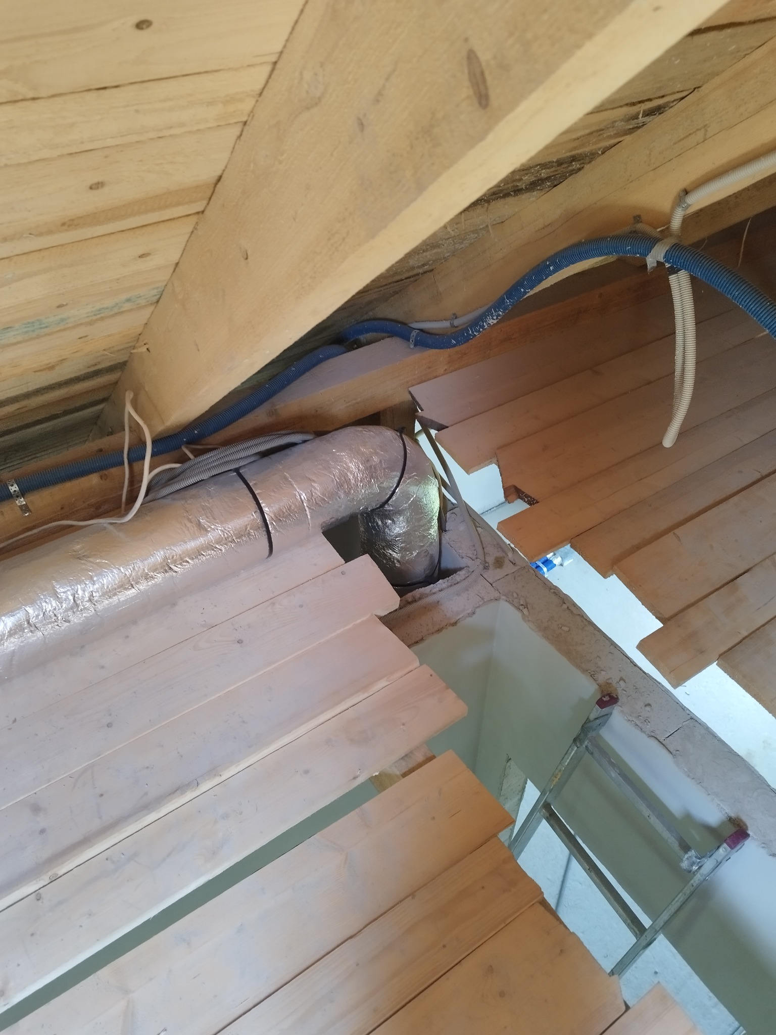 Widok z dołu na drewniany strop z zamontowanym, izolowanym kanałem wentylacyjnym, niebieskie rury instalacyjne przymocowane do belek, drabina oparta o ścianę w tle.