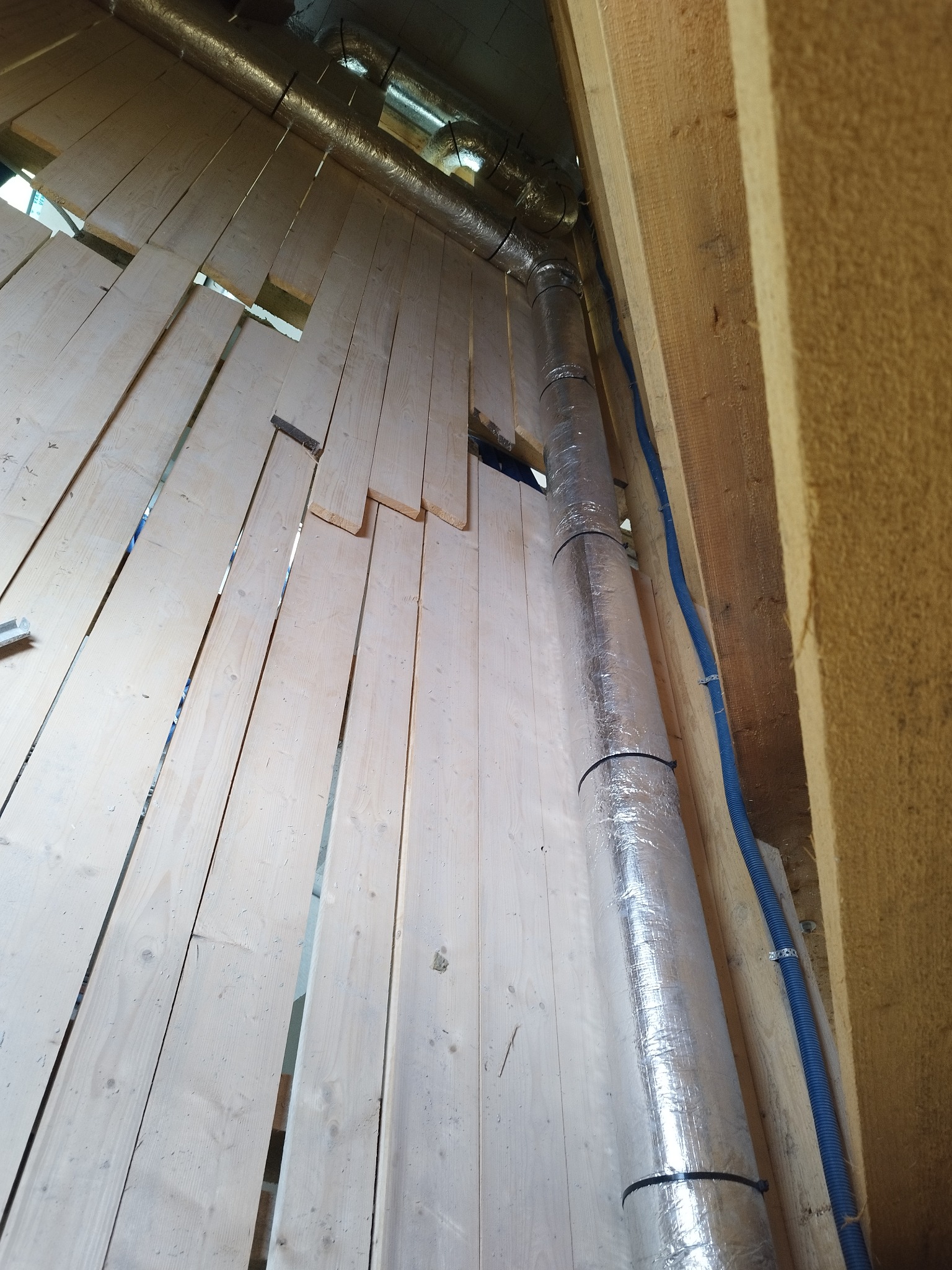 Montaż rury wentylacyjnej w przestrzeni między drewnianą ścianą a izolacją, widoczne elementy mocujące i niebieski przewód elektryczny.