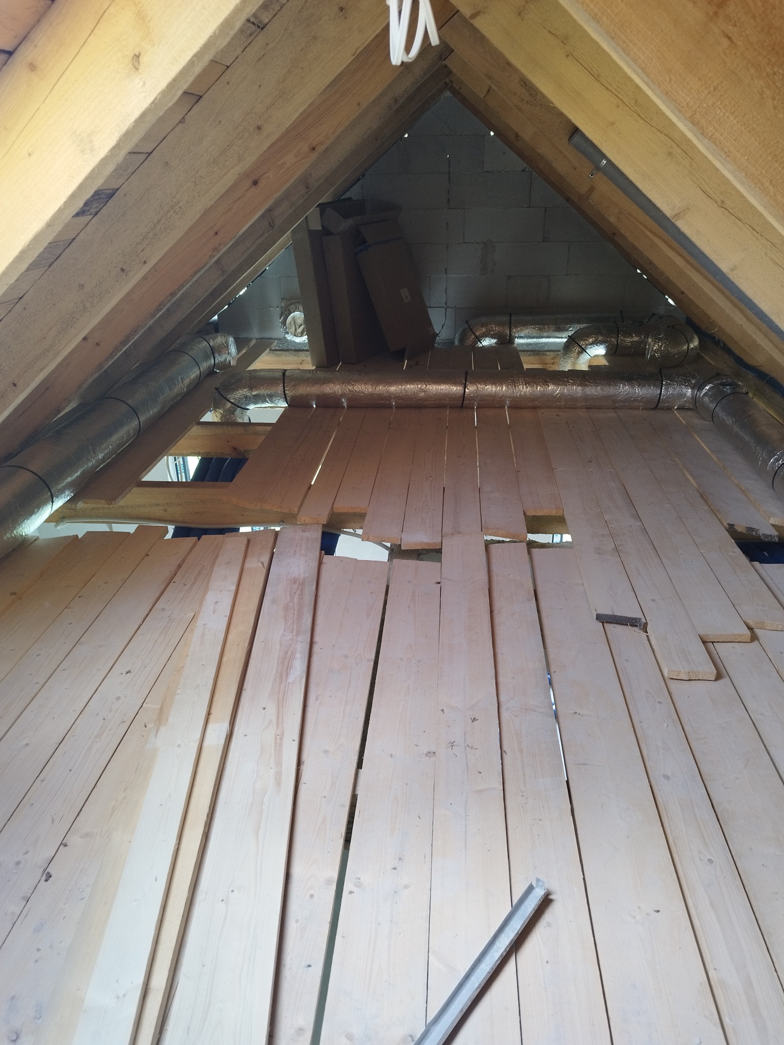 Poddasze w trakcie prac instalacyjnych: widoczne drewniane belki konstrukcyjne, częściowo ułożona podłoga z desek, srebrne rury wentylacyjne z izolacją oraz materiały budowlane składowane...