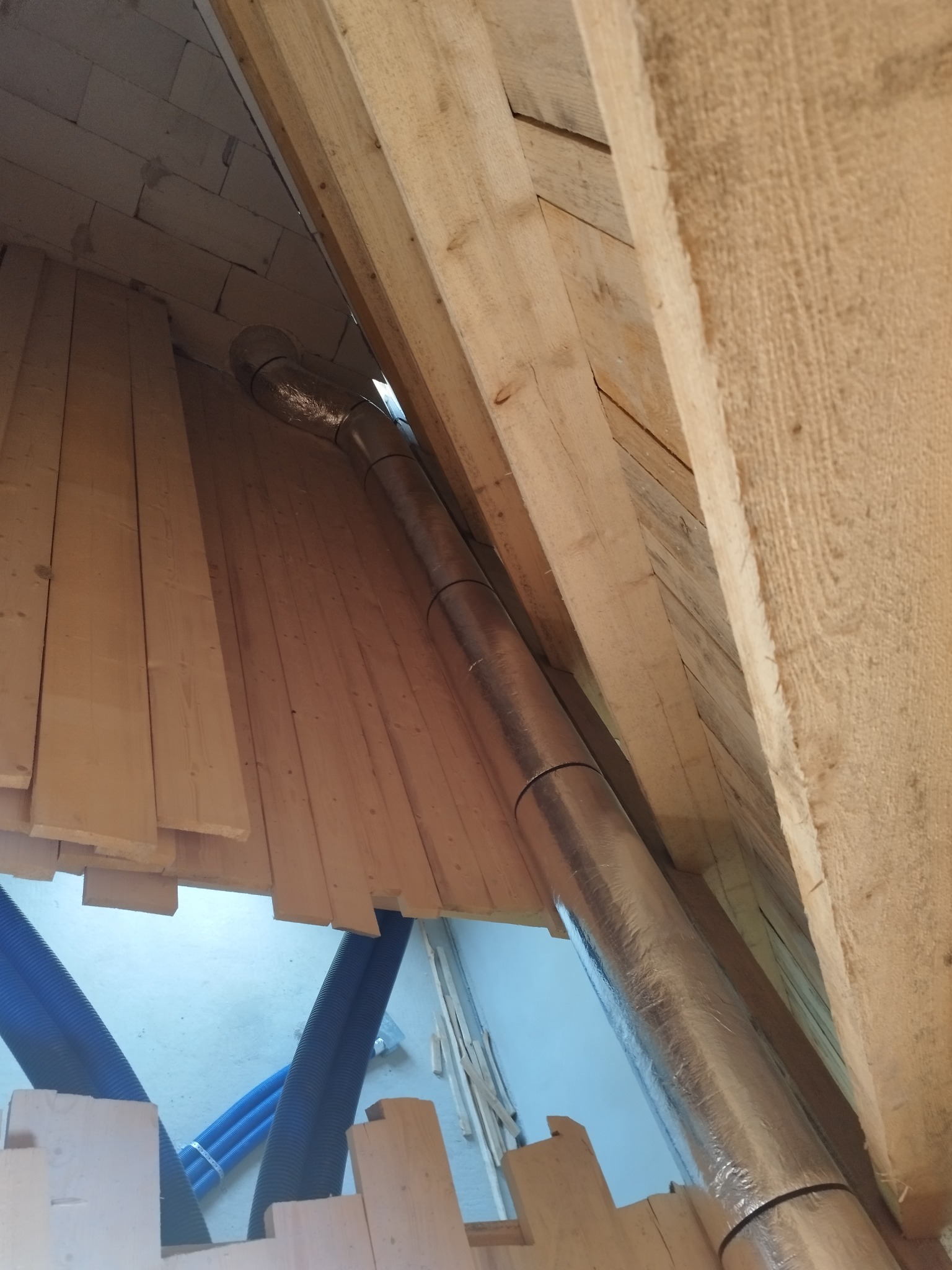Montaż izolowanego przewodu wentylacyjnego w przestrzeni poddasza z widocznymi elementami drewnianej konstrukcji dachu i niebieskimi przewodami w tle.