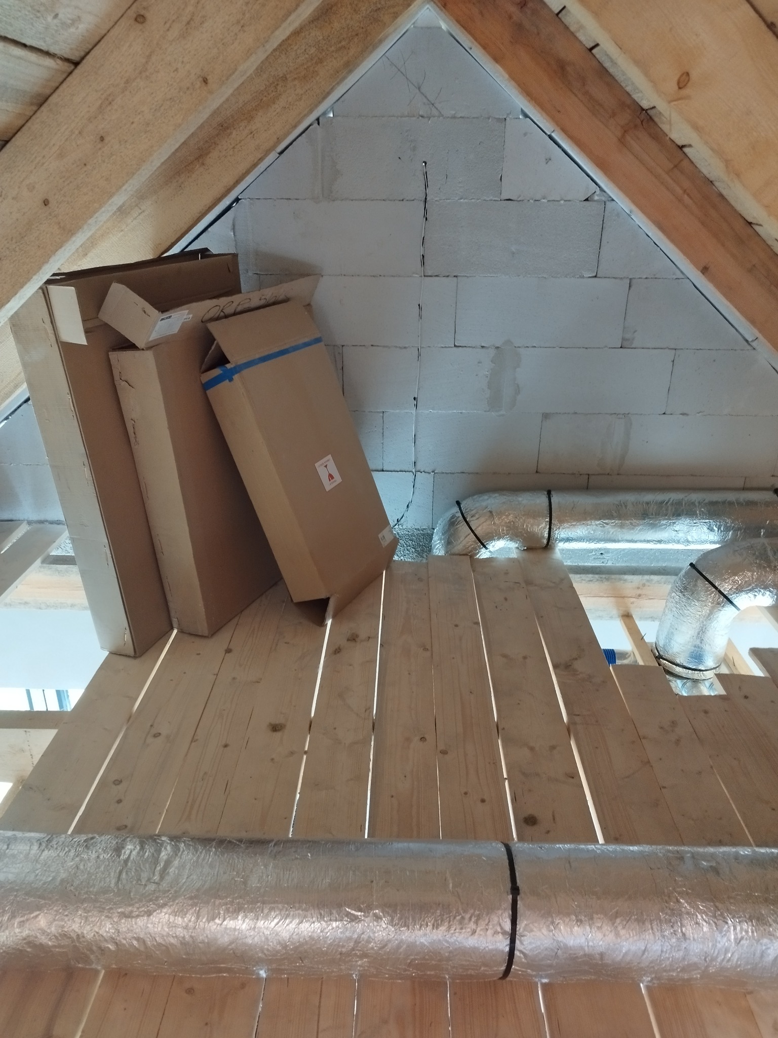 Poddasze w trakcie prac: widoczne kartony z urządzeniami, izolowane rury wentylacyjne zamocowane do drewnianej konstrukcji, ściana z betonu komórkowego.