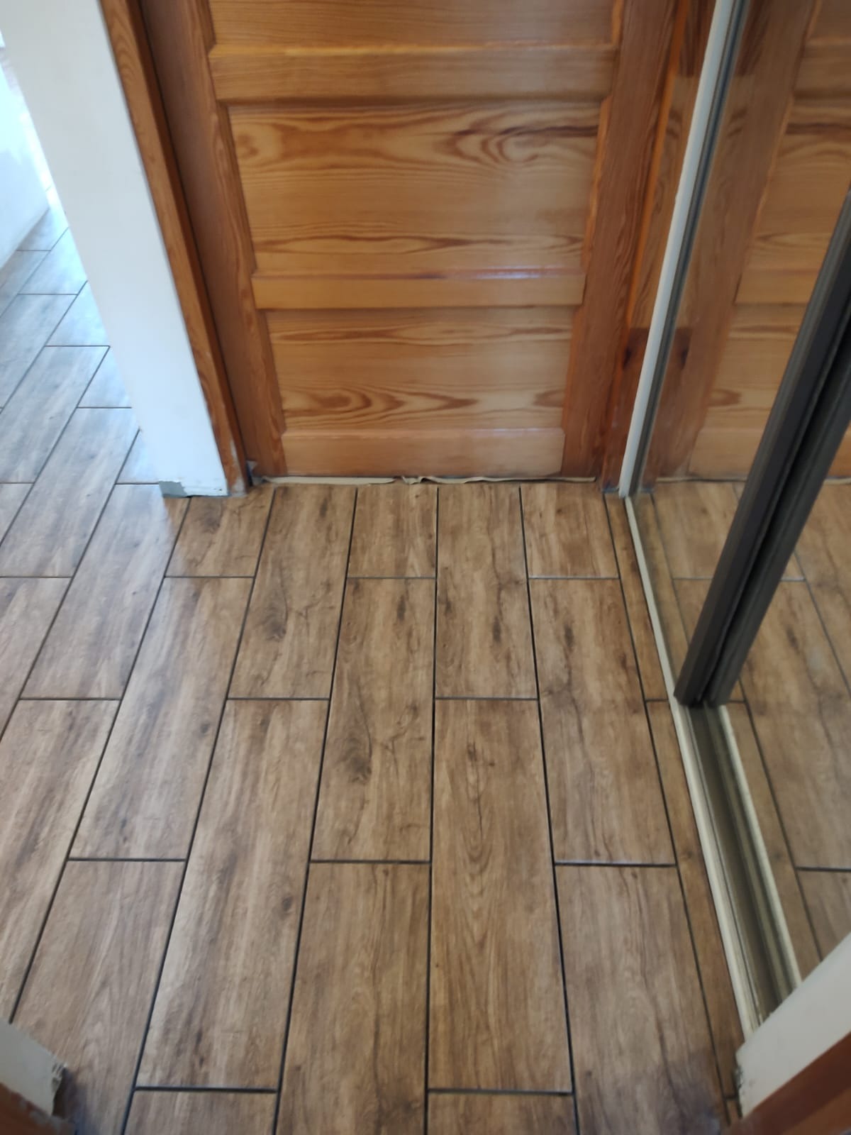 Ułożone płytki podłogowe imitujące drewno, widoczne przy drewnianych drzwiach i lustrzanych drzwiach szafy przesuwnej.