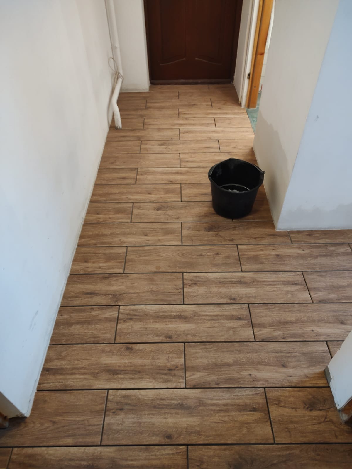 Ułożona podłoga z brązowych płytek imitujących drewno w korytarzu, widoczne białe ściany i czarne wiadro