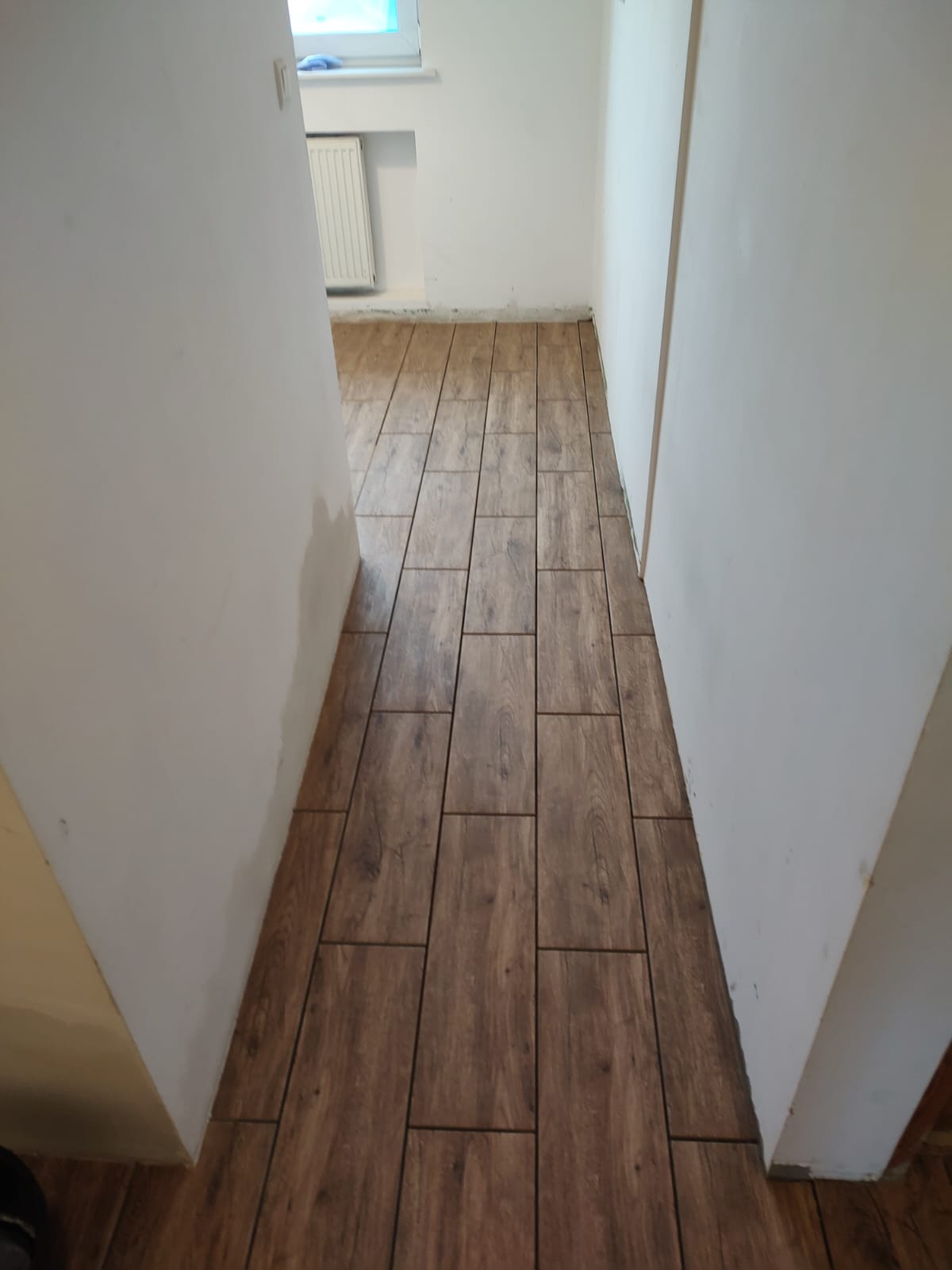 Ułożona podłoga z płytek imitujących drewno w wąskim korytarzu z białymi ścianami. Widoczny fragment okna i grzejnika.