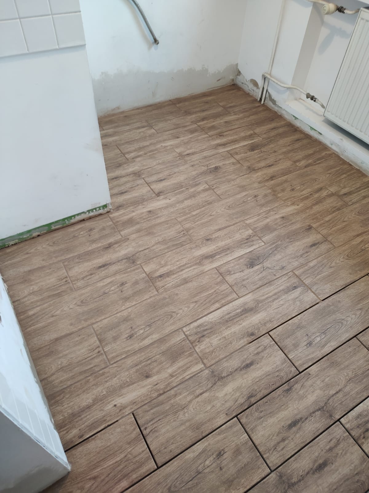 Ułożona podłoga z płytek imitujących drewno w trakcie prac wykończeniowych, widoczne niedoskonałości na ścianach i brak listew przypodłogowych.