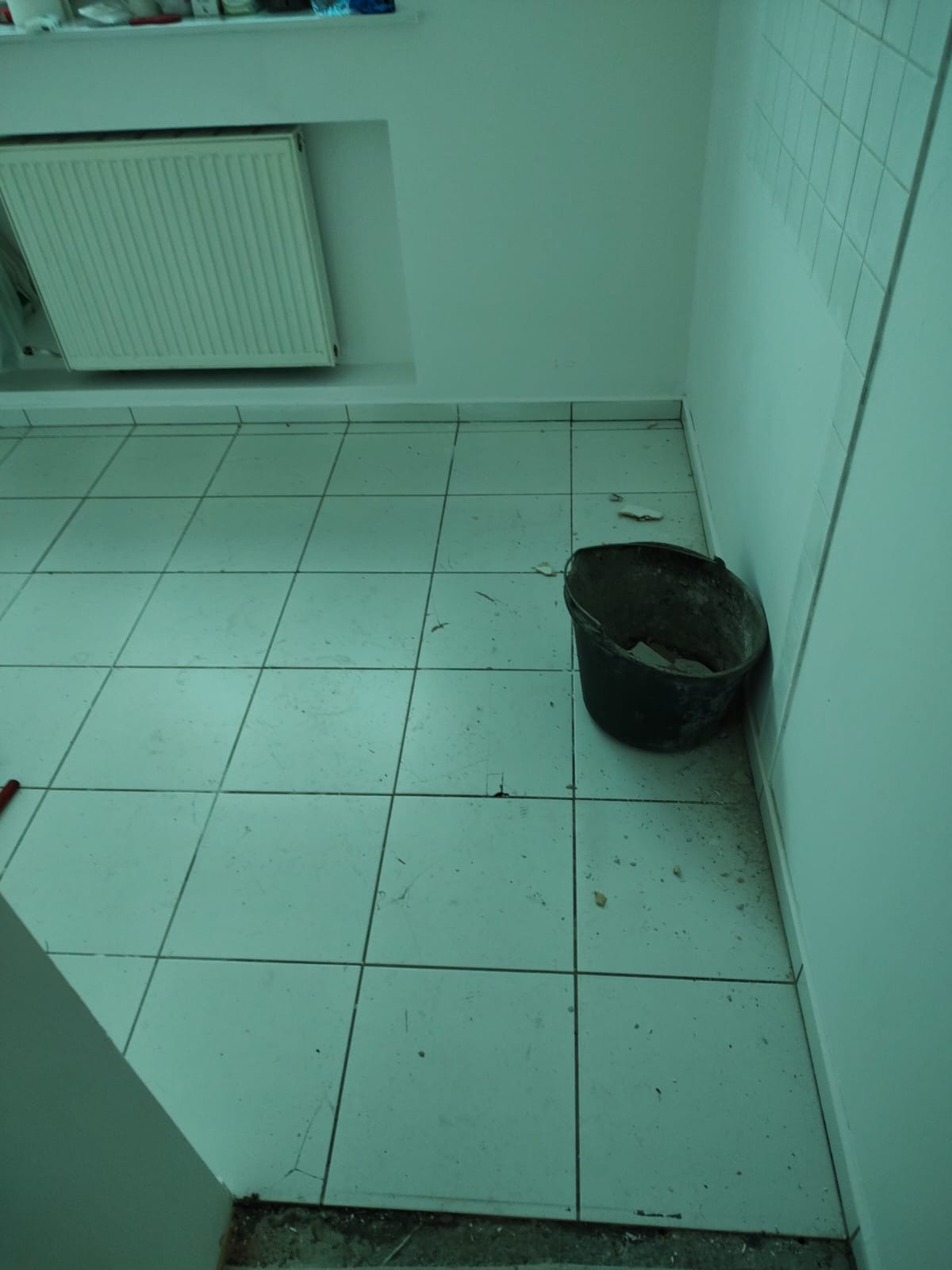 Łazienka w trakcie demontażu płytek, widoczne uszkodzone płytki podłogowe, wiadro na gruz przy ścianie, grzejnik na ścianie.
