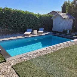 Nowoczesny basen w ogrodzie z turkusową wodą, otoczony jasnymi kamieniami i sztuczną trawą, z widoczną roletą basenową, dwoma leżakami i białą altanką w tle.