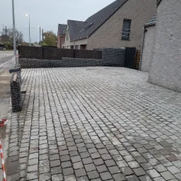 Ułożona szara kostka brukowa na podjeździe przed domem z gabionami i częściowo widocznym samochodem dostawczym z kostką w tle.
