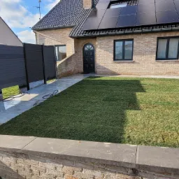 Świeżo ułożony trawnik z rolki przed domem z cegły, z czarnymi panelami słonecznymi na dachu i nowoczesnym, grafitowym ogrodzeniem panelowym po lewej stronie. Widoczny fragment kostki brukowej...