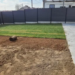 Rozwinięte rolki trawy tworzą zielony trawnik obok przygotowanego podłoża i szarej, nowoczesnej palisady ogrodzeniowej.