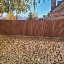 Nowoczesne ogrodzenie z pionowych desek o ciepłym, brązowym odcieniu, oddzielające posesję z brukowaną nawierzchnią pokrytą opadłymi liśćmi, z widocznym fragmentem domu z cegły w tle.