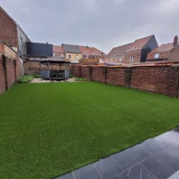 Ogród z nową, intensywnie zieloną sztuczną trawą, otoczony ceglanym murem i zabudową mieszkalną w tle, z widoczną altaną.