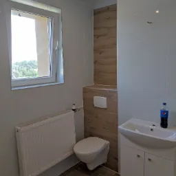 Wykończona łazienka z białymi płytkami, drewnopodobnymi akcentami, podwieszaną toaletą i białą szafką z umywalką, na której stoi butelka Pepsi.