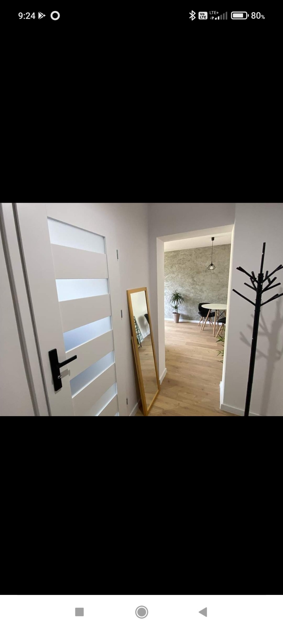 Wnętrze mieszkania z jasną podłogą, białymi drzwiami z przeszkleniami, lustrem w złotej ramie i wieszakiem na ubrania, widok na jadalnię z okrągłym stołem i rośliną doniczkową.